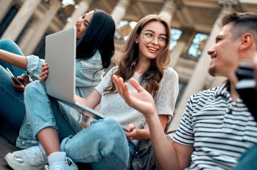 A group of students with laptops sit on the steps near the campus and communicate.