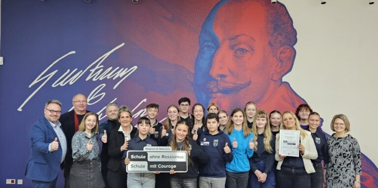 Eine Gruppe lächelnder Jugendlicher und Erwachsener vor bunter Wand mit Porträt