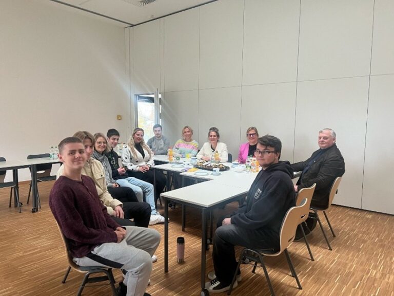 Zehn Menschen sitzen lächelnd an einem Tisch mit Tellern und Getränken im hellen Raum